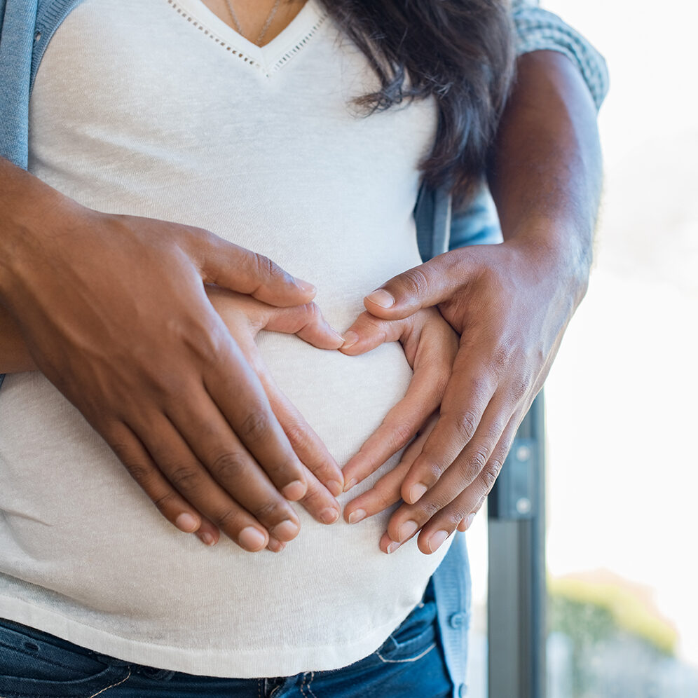 Pregnant woman and her african husband holding hand in heart shape on baby bump. Close up of multiethnic couple making heart shape on the tummy. Loving future couple expecting a baby.